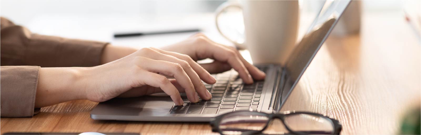 Hände von Person schreiben auf der Tastatur eines Laptops. Im Hintegrund steht ein Kaffeetasse und im Vordergrund liegt eine Brille.