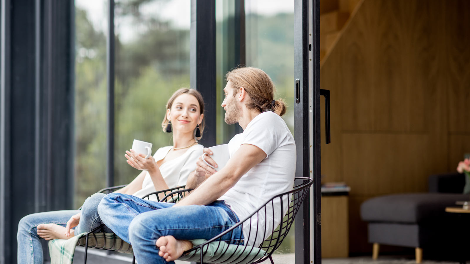 Glückliches Paar sitzt vor großen Panoramafenstern auf der Terasse mit Tassen in der Hand