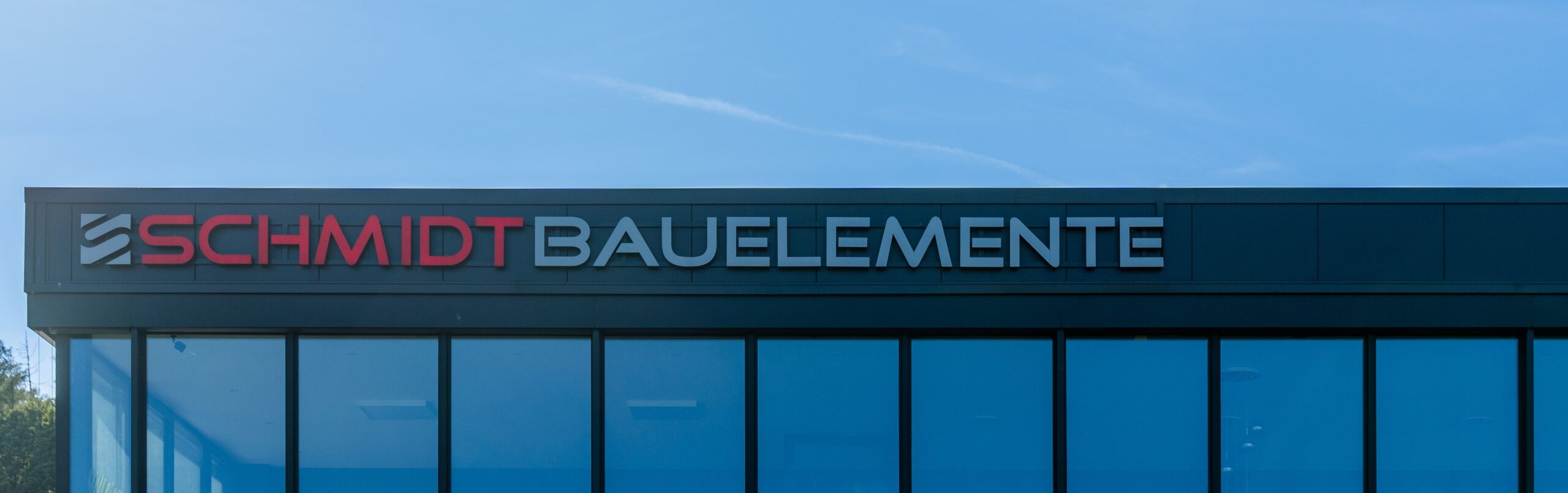 Panoramaansicht der modernen Firmenfassade von Schmidt Bauelemente mit durchgehender Glasfront und klar erkennbarer Unternehmensbeschriftung vor blauem Himmel.