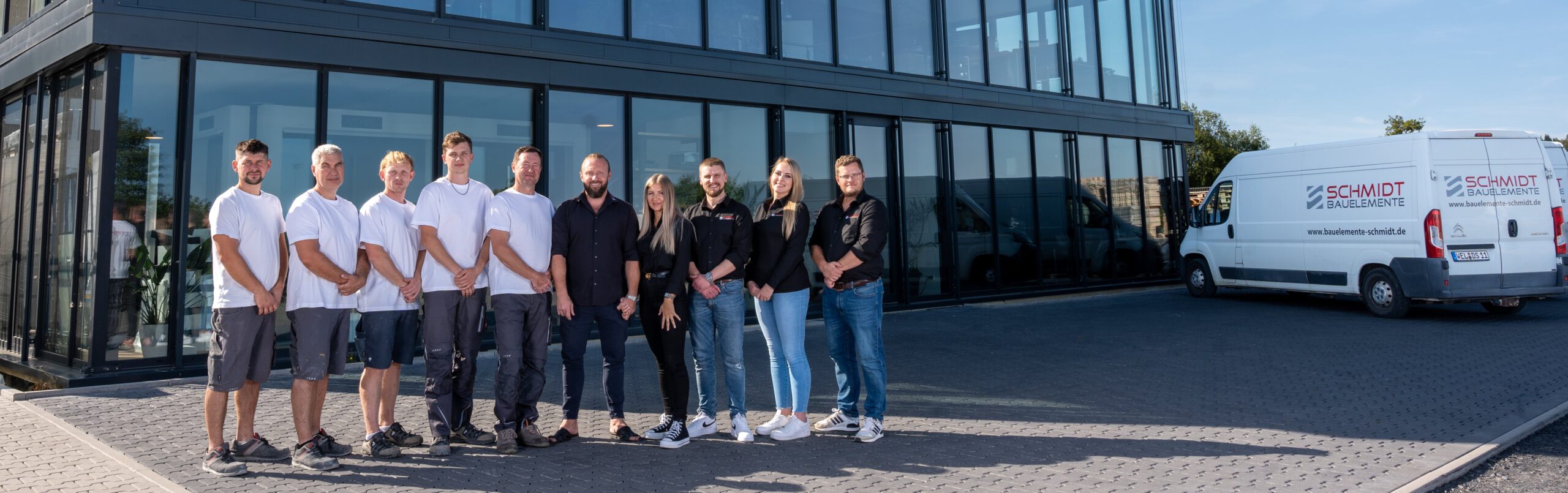 Gruppenfoto des Teams von Schmidt Bauelemente vor der modernen Firmenfassade mit Glasfront, inklusive Firmenfahrzeugen im Hintergrund.