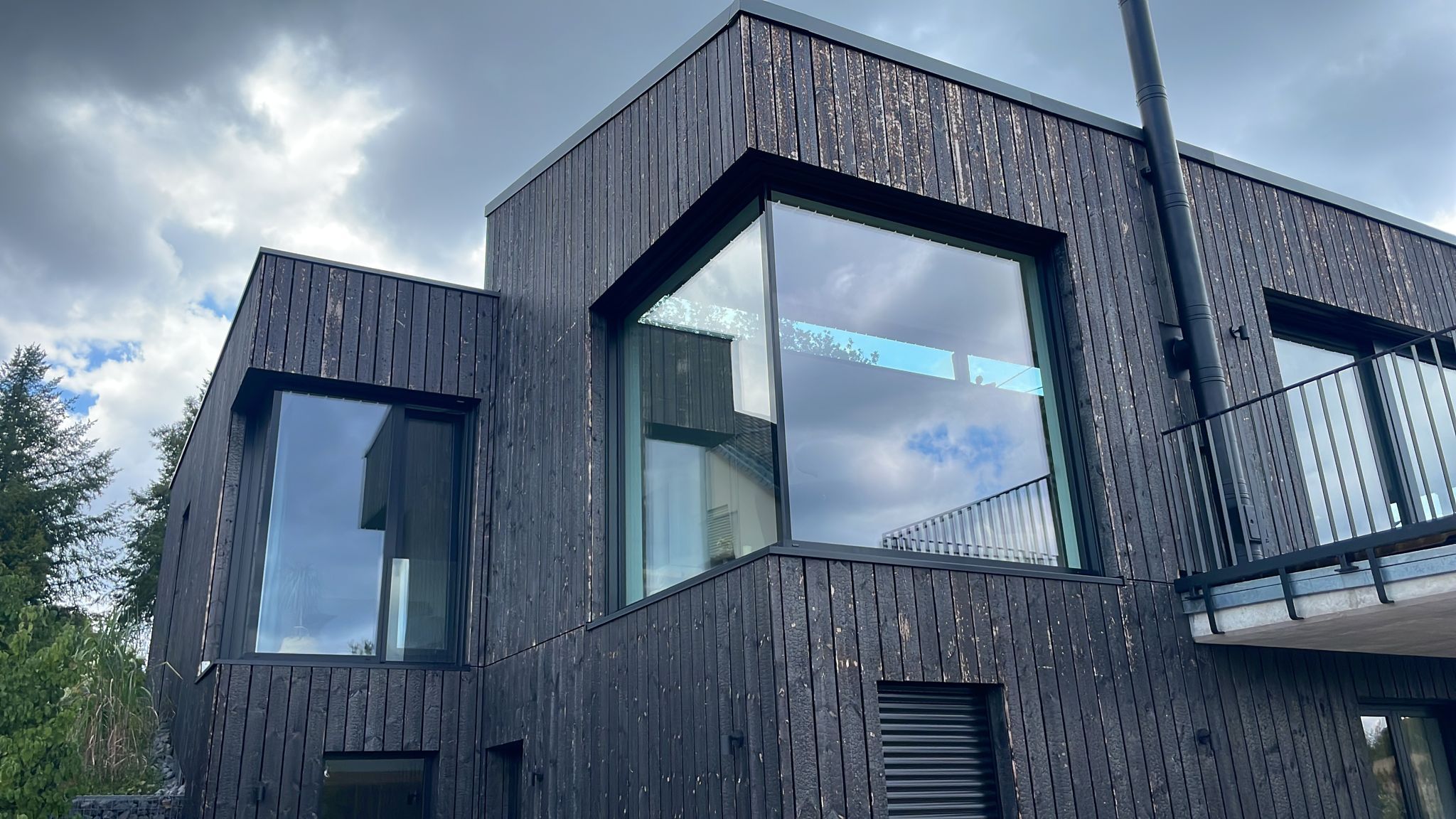 Moderne Gebäudefassade in dunkler Holzverschalung mit großzügigen, raumhohen Panoramafenstern; klare Kubatur, reduzierte Linienführung und markante Eckverglasungen, die den Innenraum optisch nach außen erweitern.