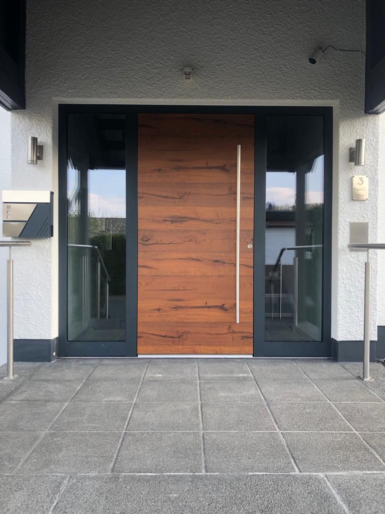 Moderne Hauseingangssituation mit einer breiten Holz-Haustür in dunklem Rahmen, flankiert von zwei bodentiefen Seitenteilen aus Glas; symmetrische Gestaltung, klarer Materialmix und reduziertes Design.