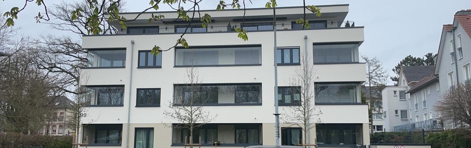 Moderne Wohnfassade mit breiten, horizontal angeordneten Fensterbändern, bodentiefen Verglasungen in den Eckbereichen und klar gerahmten Fensterformaten; durchgängige dunkelgraue Fensterprofile setzen präzise Kontraste zur hellen Putzfläche und betonen die lineare Gebäudearchitektur.