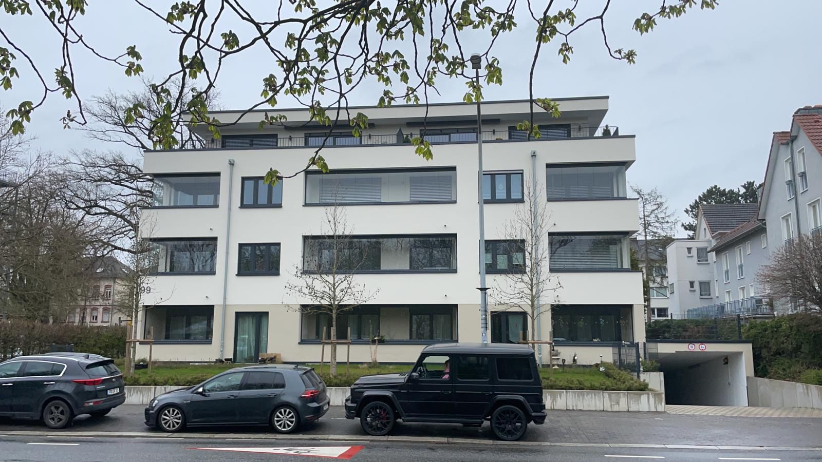 Moderne Wohnfassade mit breiten, horizontal angeordneten Fensterbändern, bodentiefen Verglasungen in den Eckbereichen und klar gerahmten Fensterformaten; durchgängige dunkelgraue Fensterprofile setzen präzise Kontraste zur hellen Putzfläche und betonen die lineare Gebäudearchitektur.