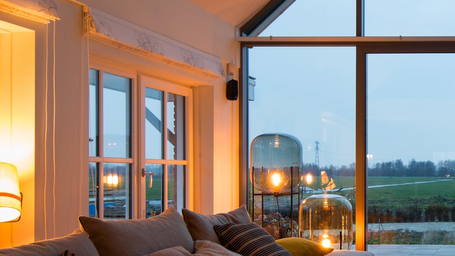 Modernes Wohnzimmer mit großen Fensterfronten, Sofa und warmem Licht mit Blick auf eine ländliche Abendlandschaft.
