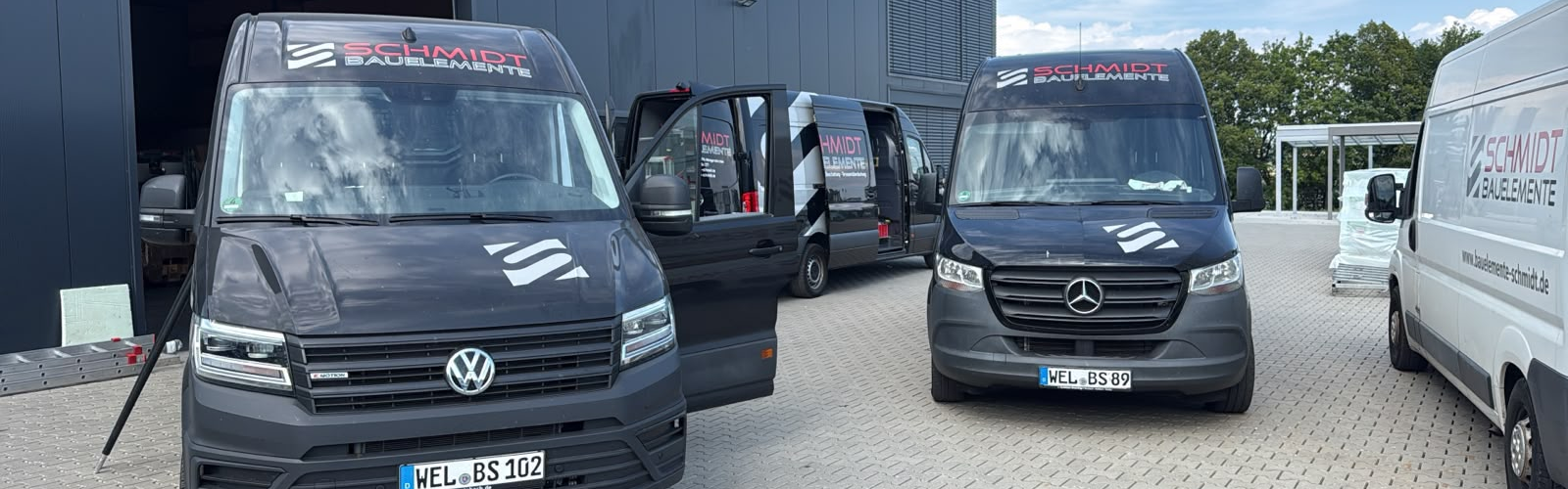 Mehrere beschriftete Lieferwagen von Schmidt Bauelemente stehen vor einer Gewerbehalle, darunter ein VW- und ein Mercedes-Transporter.